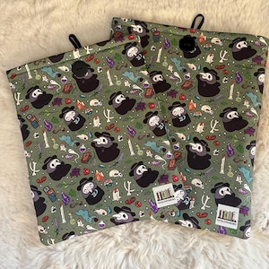 Op de afbeelding: Twee groene stoffen zakjes met een zwarte knoopsluiting. De zakjes hebben een herhalend patroon van een pestdokter met een masker, schedels, kristallen en andere occulte afbeeldingen. De tekst "CatsLoveBooks" staat gedrukt op de onderkant van elk zakje.