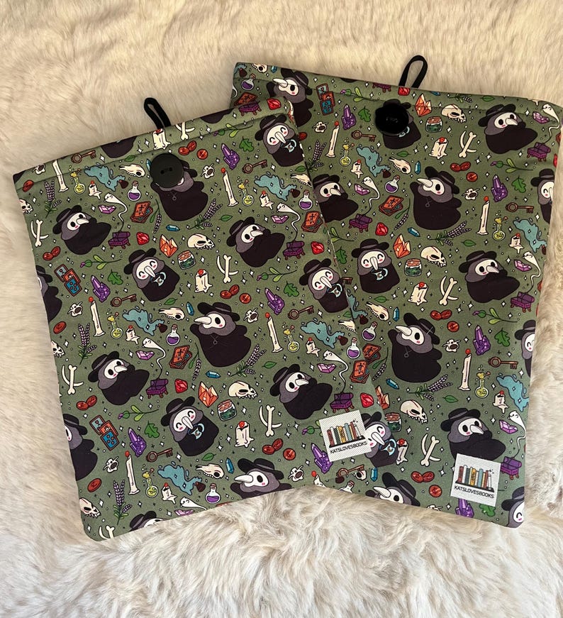 Op de afbeelding: Twee groene stoffen zakjes met een zwarte knoopsluiting. De zakjes hebben een herhalend patroon van een pestdokter met een masker, schedels, kristallen en andere occulte afbeeldingen. De tekst "CatsLoveBooks" staat gedrukt op de onderkant van elk zakje.