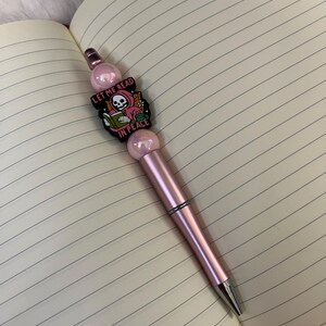 May include: A pink pen with a charm featuring a skeleton reading a book with the text "Let me read in peace".