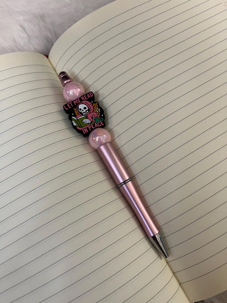 May include: A pink pen with a charm featuring a skeleton reading a book with the text "Let me read in peace".