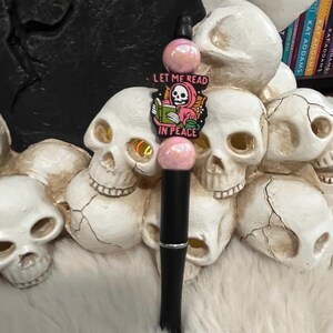 May include: A black pen with pink and white beads and a charm that says "Let me read in peace" with a cartoon skeleton reading a book.
