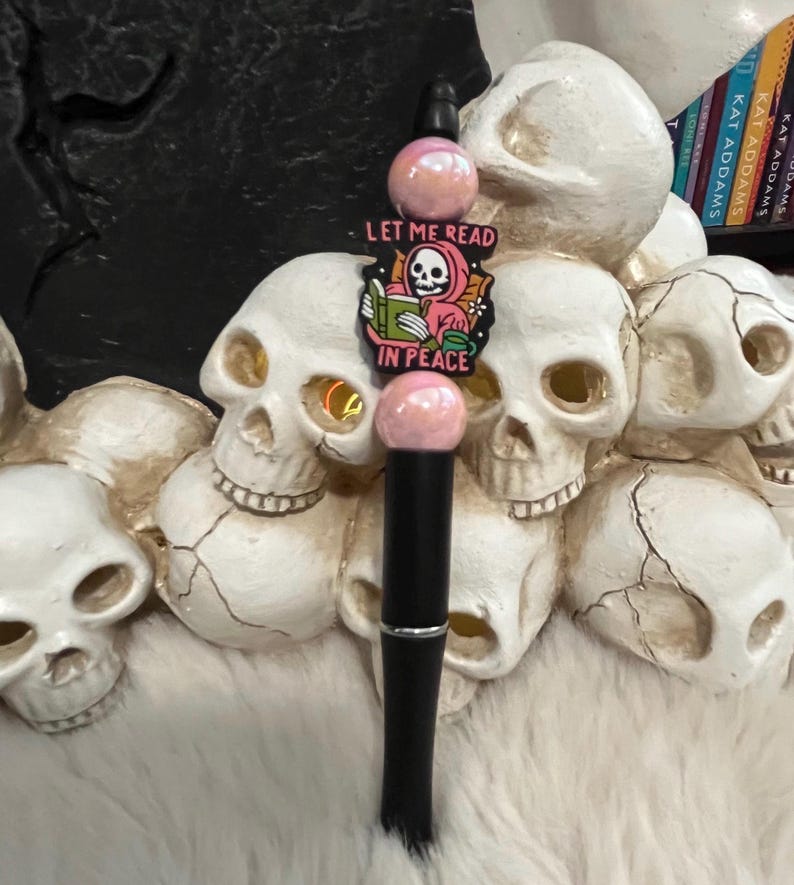 May include: A black pen with pink and white beads and a charm that says "Let me read in peace" with a cartoon skeleton reading a book.