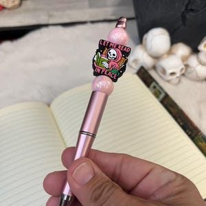 May include: A pink pen with a black and white enamel charm featuring a skeleton reading a book. The charm says "Let me read in peace".