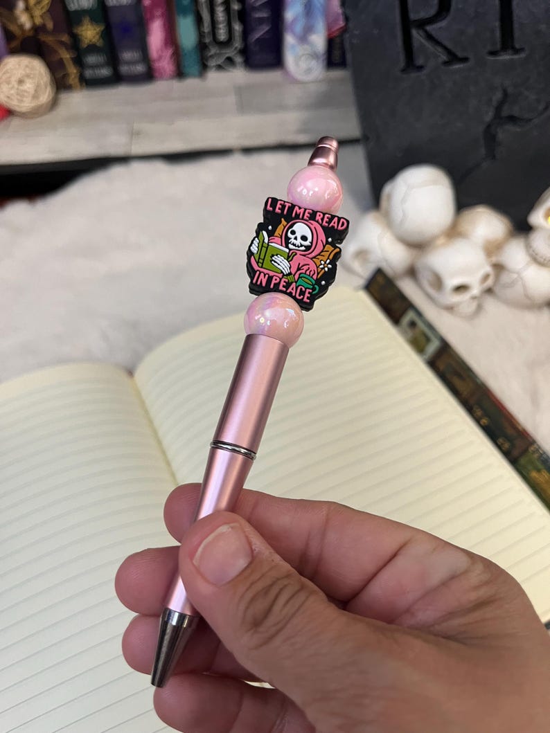 May include: A pink pen with a black and white enamel charm featuring a skeleton reading a book. The charm says "Let me read in peace".