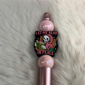 May include: A pink and rose gold pen with a black charm featuring a skeleton reading a book and the text "Let me read in peace".