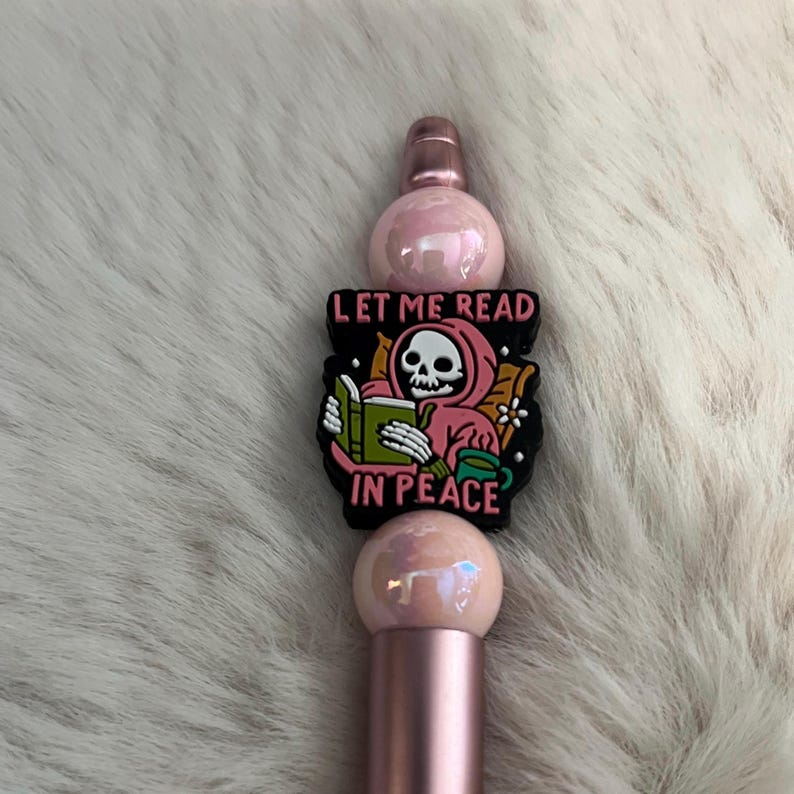 May include: A pink and rose gold pen with a black charm featuring a skeleton reading a book and the text "Let me read in peace".