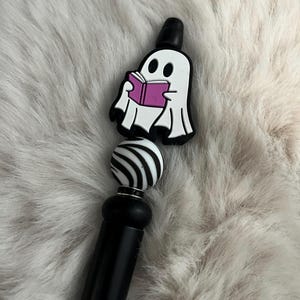 May include: A black and white striped bead with a black base and a white ghost charm holding a pink book. The ghost charm is attached to the top of the bead.