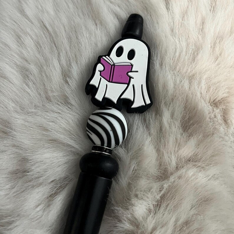 May include: A black and white striped bead with a black base and a white ghost charm holding a pink book. The ghost charm is attached to the top of the bead.