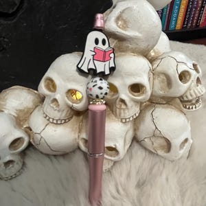 May include: A pink and gold pen with a white ghost charm holding a pink book. The charm is attached to a black and white speckled bead. The pen is surrounded by white ceramic skulls.