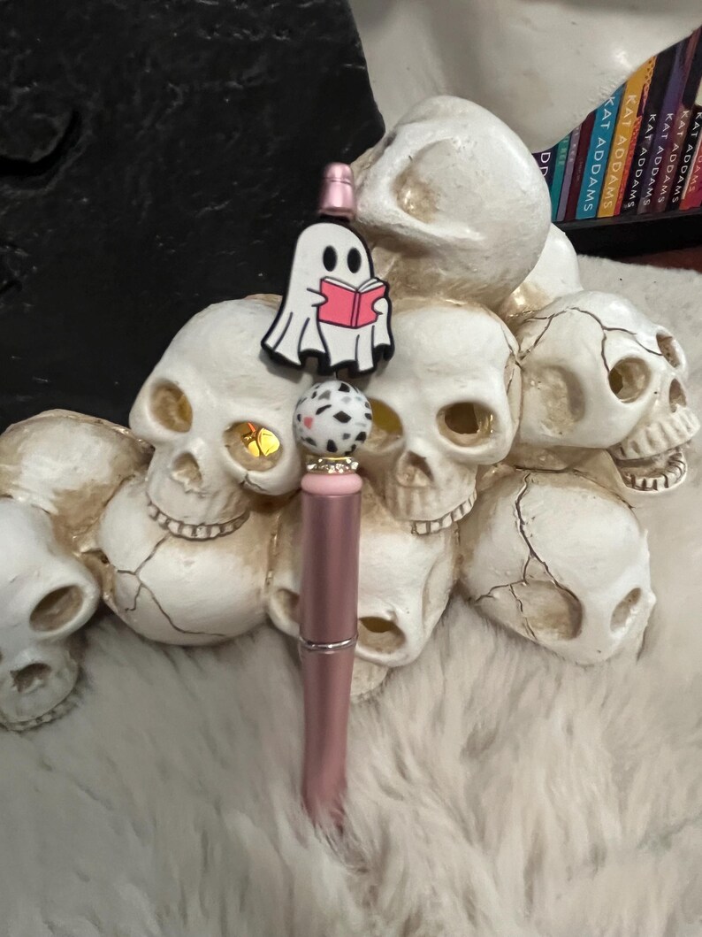 May include: A pink and gold pen with a white ghost charm holding a pink book. The charm is attached to a black and white speckled bead. The pen is surrounded by white ceramic skulls.