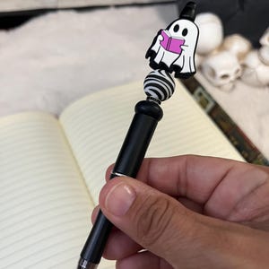 May include: A black pen with a white and black striped bead and a ghost reading a pink book charm. The pen is held in a hand.