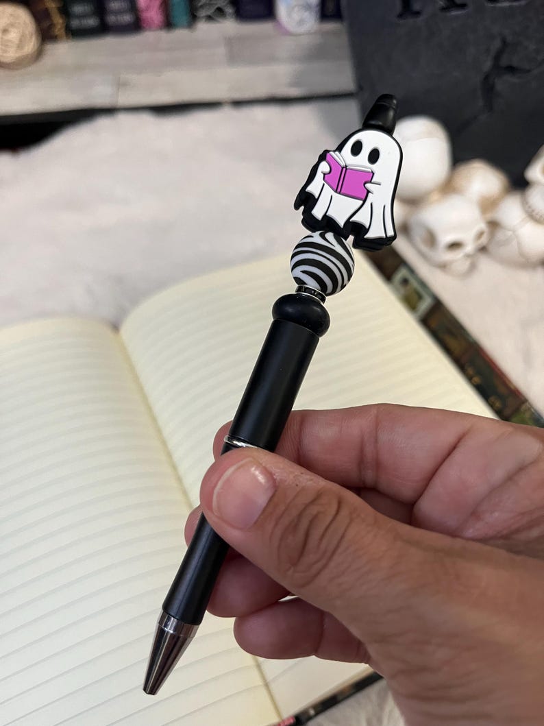 May include: A black pen with a white and black striped bead and a ghost reading a pink book charm. The pen is held in a hand.