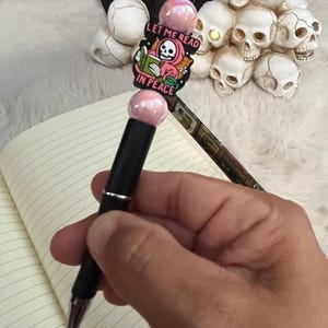 May include: A black pen with a pink and white charm that says "Let me read in peace". The charm features a skeleton wearing a pink hooded robe and holding a book.
