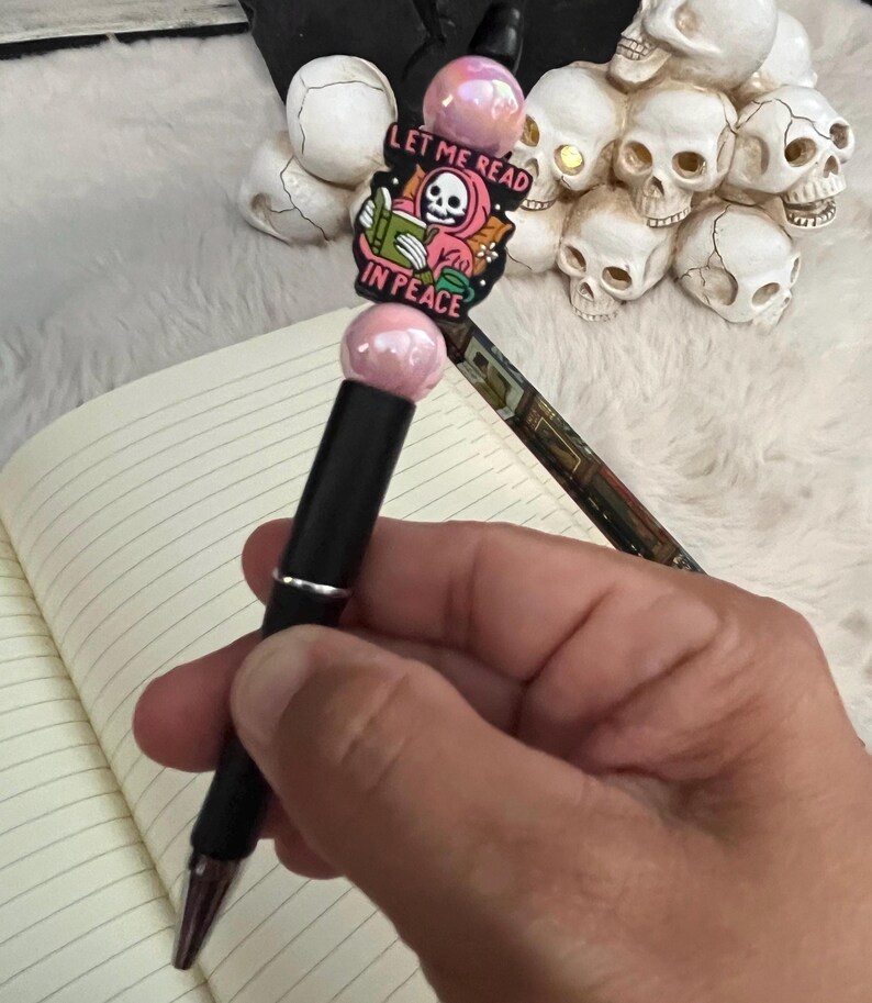 May include: A black pen with a pink and white charm that says "Let me read in peace". The charm features a skeleton wearing a pink hooded robe and holding a book.