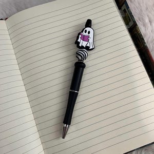 May include: A black pen with a white and black striped bead and a ghost holding a book charm. The ghost is pink and white.
