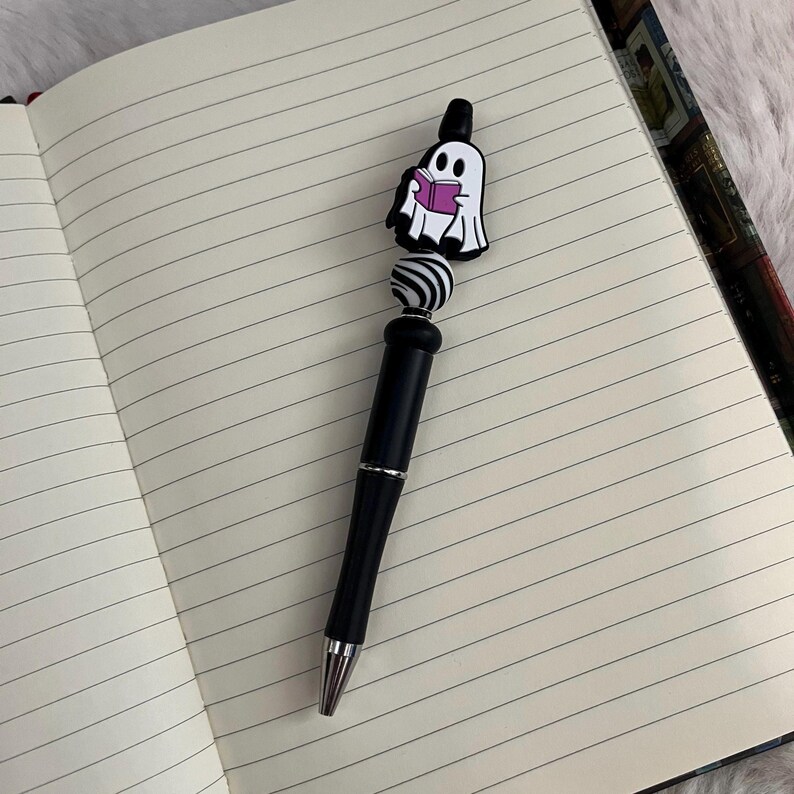 May include: A black pen with a white and black striped bead and a ghost holding a book charm. The ghost is pink and white.