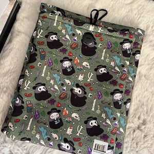 Op de afbeelding: Groene trekkoordtas met een herhalend patroon van pestdokters, schedels, botten, kristallen en andere benodigdheden voor het maken van drankjes. De tas heeft een zwarte knoopsluiting en de tekst "KATSLOVESBOOKS" is onderaan gedrukt.