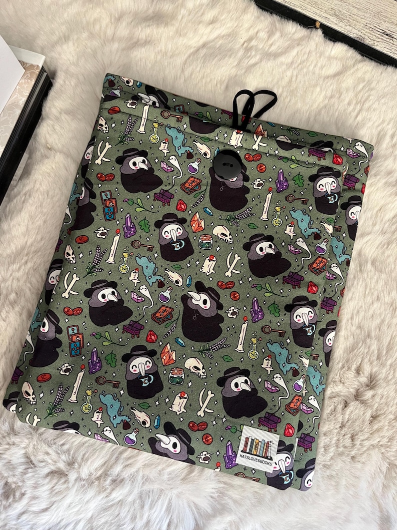 Op de afbeelding: Groene trekkoordtas met een herhalend patroon van pestdokters, schedels, botten, kristallen en andere benodigdheden voor het maken van drankjes. De tas heeft een zwarte knoopsluiting en de tekst "KATSLOVESBOOKS" is onderaan gedrukt.