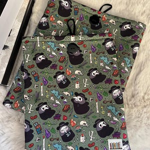 Op de afbeelding: Twee groene stoffen zakken met een trekkoord. De zakken hebben een herhalend patroon van pestdokters met snavelmaskers, schedels, botten, drankflesjes en andere griezelige elementen. De tekst "KATSLOVESBOOKS" staat gedrukt in de rechterbenedenhoek van de zak.