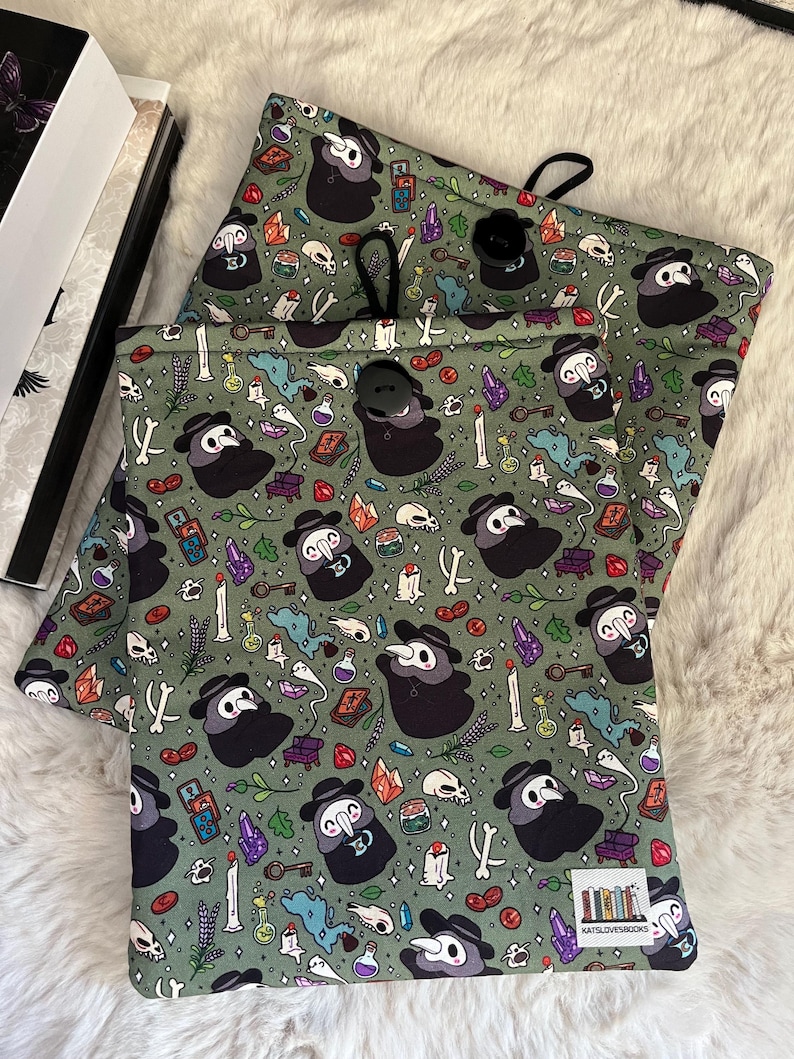 Op de afbeelding: Twee groene stoffen zakken met een trekkoord. De zakken hebben een herhalend patroon van pestdokters met snavelmaskers, schedels, botten, drankflesjes en andere griezelige elementen. De tekst "KATSLOVESBOOKS" staat gedrukt in de rechterbenedenhoek van de zak.