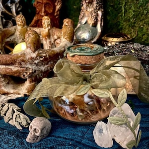 Pode incluir: Um pequeno frasco de vidro cheio de ervas e flores secas, amarrado com uma fita verde. Uma pequena caveira, cristais de quartzo e estátuas decorativas são dispostos ao redor do frasco. O fundo apresenta musgo e outros elementos decorativos.