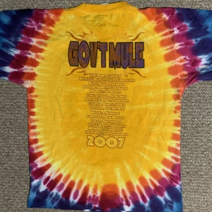 Può includere: Una maglietta gialla tie-dye con il nome della band "GOV'T MULE" stampato in marrone sul davanti. La maglietta elenca anche le date del tour della band del 2007 in testo nero.