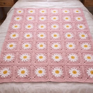 Op de afbeelding: Een roze gehaakte deken met een madeliefjespatroon. De deken heeft witte en gele madeliefjesmotieven. De deken ligt op een bed met witte lakens en kussens. De deken heeft een vierkante vorm.