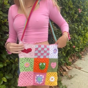 May include: A colorful crochet shoulder bag with a patchwork design. The bag features squares in various colors, including pink, green, orange, and white, with heart, flower, and checkerboard patterns. The strap is rainbow-colored.
