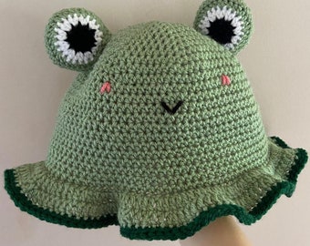 May include: A light green crochet bucket hat featuring two protruding frog eyes with white and black details. The hat has a wide, ruffled brim with a dark green edge, and embroidered pink cheeks and a black V-shaped mouth on the front.
