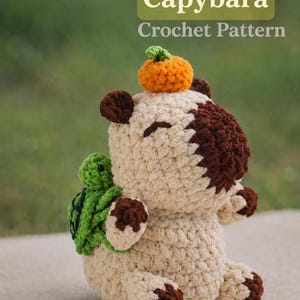 May include: A beige and brown crocheted capybara toy with a small orange pumpkin on its head and a tiny green turtle attached to its back. The text Capybara Crochet Pattern is displayed in white and light green above the stuffed animal figure.