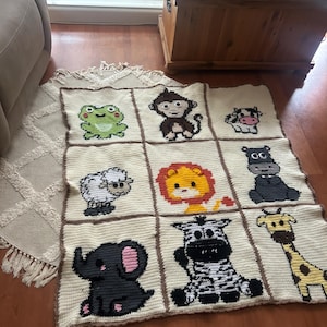 Könnte beinhalten: Eine gehäkelte Babydecke mit weißem Hintergrund und braunen Quadraten. Jedes Quadrat zeigt ein anderes Tier, darunter ein Frosch, ein Affe, eine Kuh, ein Flusspferd, ein Schaf, ein Löwe, ein Zebra, eine Giraffe und ein Elefant.