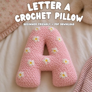 May include: A pink crochet pillow shaped like the letter A, featuring white daisy appliques with yellow centers. The text above reads LETTER A CROCHET PILLOW BEGINNER FRIENDLY + PDF DOWNLOAD. The pillow has a textured, yarn-based surface.