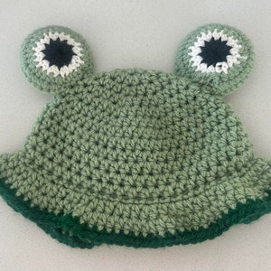 Puede incluir: Sombrero de rana de crochet verde con ojos blancos y negros. El sombrero tiene un borde verde con un ribete verde oscuro.