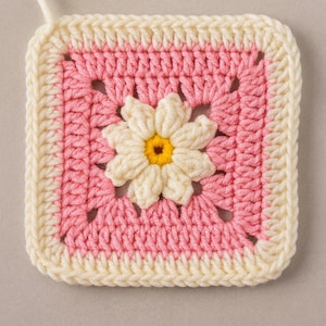 Könnte beinhalten: Ein quadratisches, handgefertigtes Häkelstück mit einem Gänseblümchen-Design. Das Quadrat hat eine rosa Mitte mit einer cremefarbenen Gänseblümchen in der Mitte und einen cremefarbenen Rand. Das Gänseblümchen hat eine gelbe Mitte.