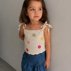 May include: A cream-colored crochet top featuring thin tie-shoulder straps and a scalloped hem. The garment is decorated with small, colorful floral appliques in shades of pink, yellow, orange, and green. The text Crochet Kids Top Pattern is visible.