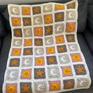 Puede incluir: Una manta de bebé tejida a crochet con un patrón repetitivo de soles, lunas y estrellas. La manta está hecha en tonos de blanco, rosa, marrón y amarillo.