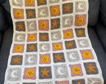 May include: A crocheted baby blanket with a repeating pattern of suns, moons, and stars. The blanket is made in shades of white, pink, brown, and yellow.