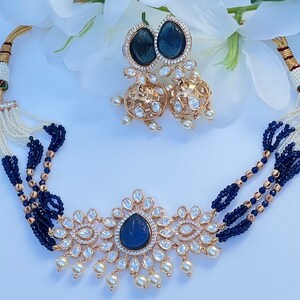 Peut inclure: Un collier en or avec un pendentif en pierre bleue et des accents de perles blanches. Le collier est composé de plusieurs rangs de perles bleues et blanches. Le pendentif est entouré de pierres plus petites. Le collier est accompagné de boucles d'oreilles assorties avec des pierres bleues et des perles blanches.