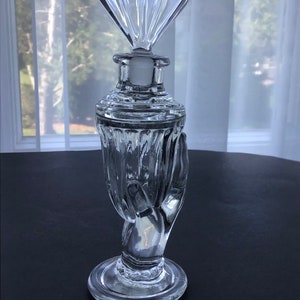 Peut inclure: Une bouteille en verre transparent avec une base en forme de main et un bouchon en forme d'éventail. La bouteille est décorée de lignes verticales.