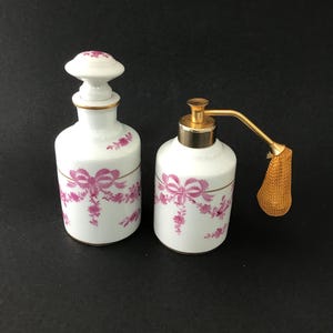 May include: Two white porcelain vanity bottles featuring a pink floral and ribbon pattern with gold trim. One bottle has a matching stopper, while the other features a gold-toned metal pump mechanism with an attached gold mesh atomizer bulb.