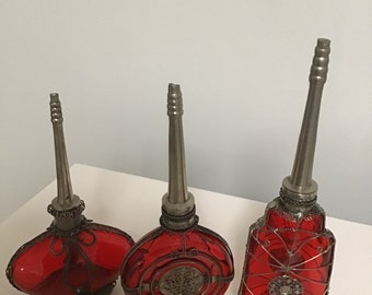 May include: Three red glass bottles with silver metal tops and intricate wire designs. The bottles have a round, oval, and rectangular shape.