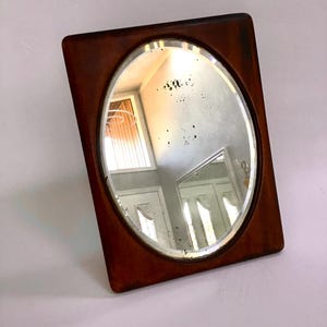 May include: A vintage mirror with an oval reflective surface set in a dark brown, rectangular frame. The mirror reflects a room with white doors and a window with blinds. The frame has rounded corners.