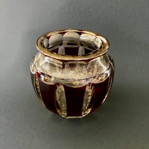 Puede incluir: Un pequeño jarrón de cristal transparente con cuerpo redondeado y boca ancha. El jarrón presenta un color rojo intenso en la parte inferior y un borde dorado. Intrincados diseños dorados adornan la sección superior, añadiendo un toque de elegancia. El jarrón es probablemente decorativo.