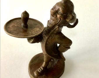 May include: A bronze-toned metal figurine of a smiling person in period clothing, standing on a circular base. The figure holds a round tray in one hand, which features a central upright handle and engraved text along the outer rim.