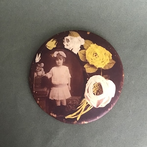 Puede incluir: Un espejo redondo, marrón y decorativo con un diseño floral blanco y amarillo. Una niña pequeña con un vestido blanco aparece en el centro del espejo.