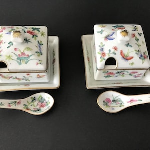 以下が含まれることがあります： ふた付きの白い磁器製の調味料セット2つと、それに合うスプーン2本。それぞれのセットには、蝶と金色の縁取りが施された花柄が描かれています。スプーンにも同じような花柄が描かれています。