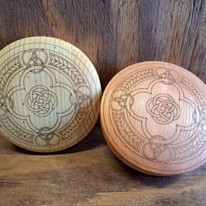May include: Two round wooden objects with intricate Celtic knot designs carved into their surfaces. One is light-colored, the other a reddish-brown. The designs include interwoven knots and leaf patterns, creating a decorative effect.