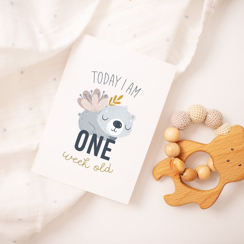 May include: A baby milestone card with a cartoon bear illustration and the text "TODAY I AM ONE week old." A wooden baby rattle with a bear-shaped handle and wooden beads is also in the image. The card and rattle are on a white surface.