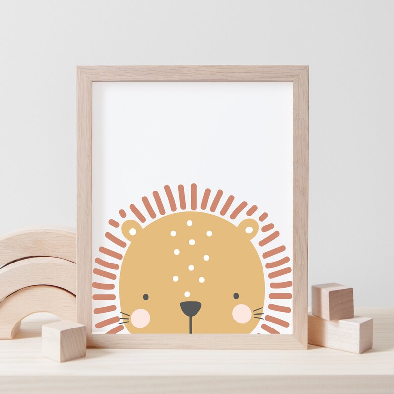 May include: A framed print of a cartoon lion's face. The lion has a tan face, a dark grey nose, pink cheeks, and a rust-coloured mane. The print is in a light-coloured wooden frame, suitable for a nursery or children's room.