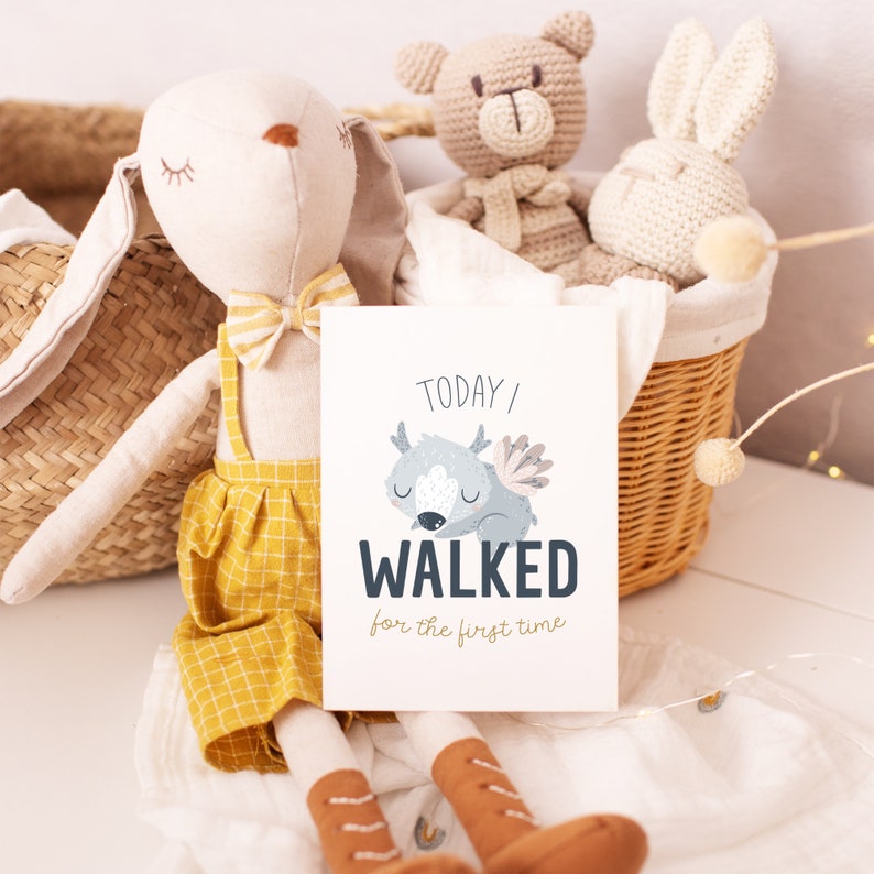 May include: A stuffed bunny doll with a yellow bow tie and overalls sits next to a card that reads "Today I walked for the first time." A woven basket holds a crocheted bear and bunny. The scene is soft and inviting, perfect for a nursery.